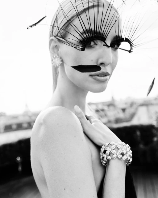 Black and white portrait of a fashionable woman with large decorative eyelashes, statement earrings, and a jeweled bracelet outdoors.