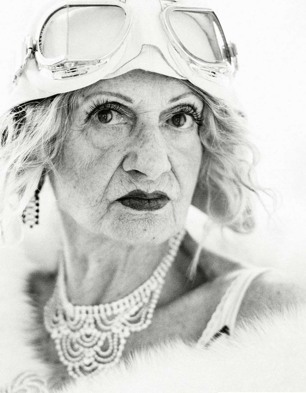 Monochrome portrait of an older woman wearing aviator goggles and vintage jewelry, creating a retro or historical fashion statement.