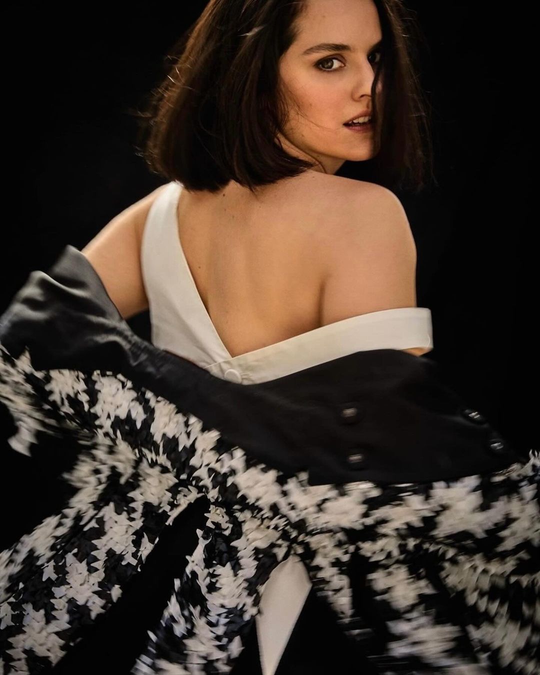 Brunette woman in white dress and patterned jacket poses in a studio with a black background, looking over her shoulder.