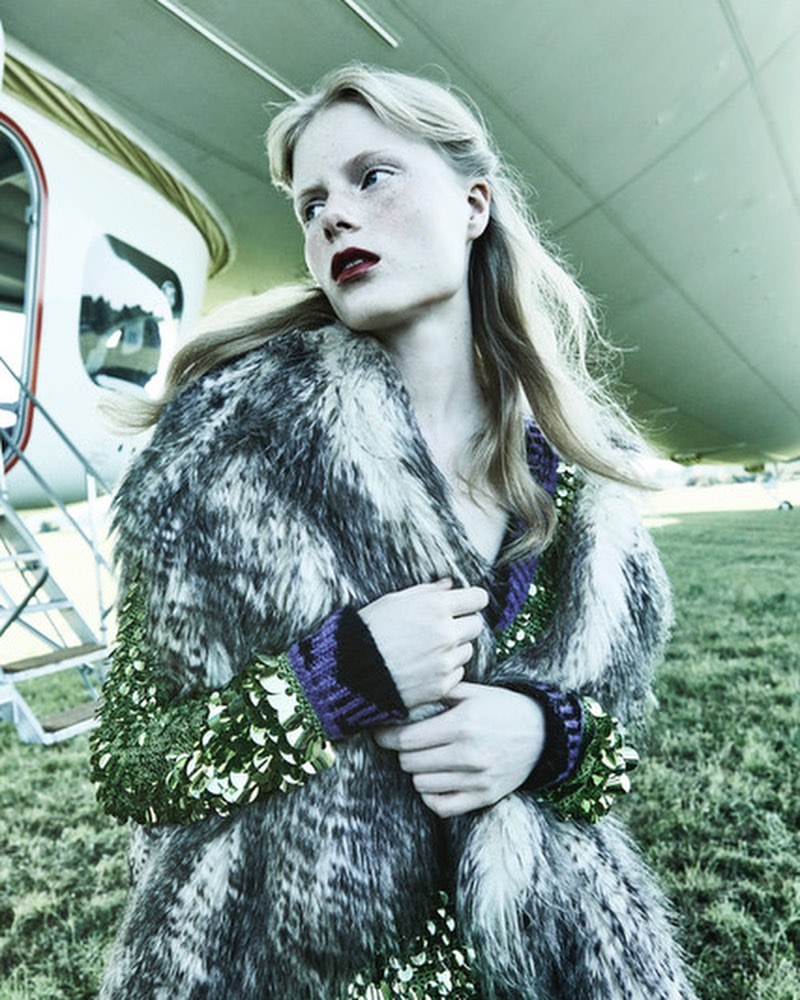 Woman in a fur coat poses outdoors near a plane. The image captures a fashion shot with the woman as the focal point.