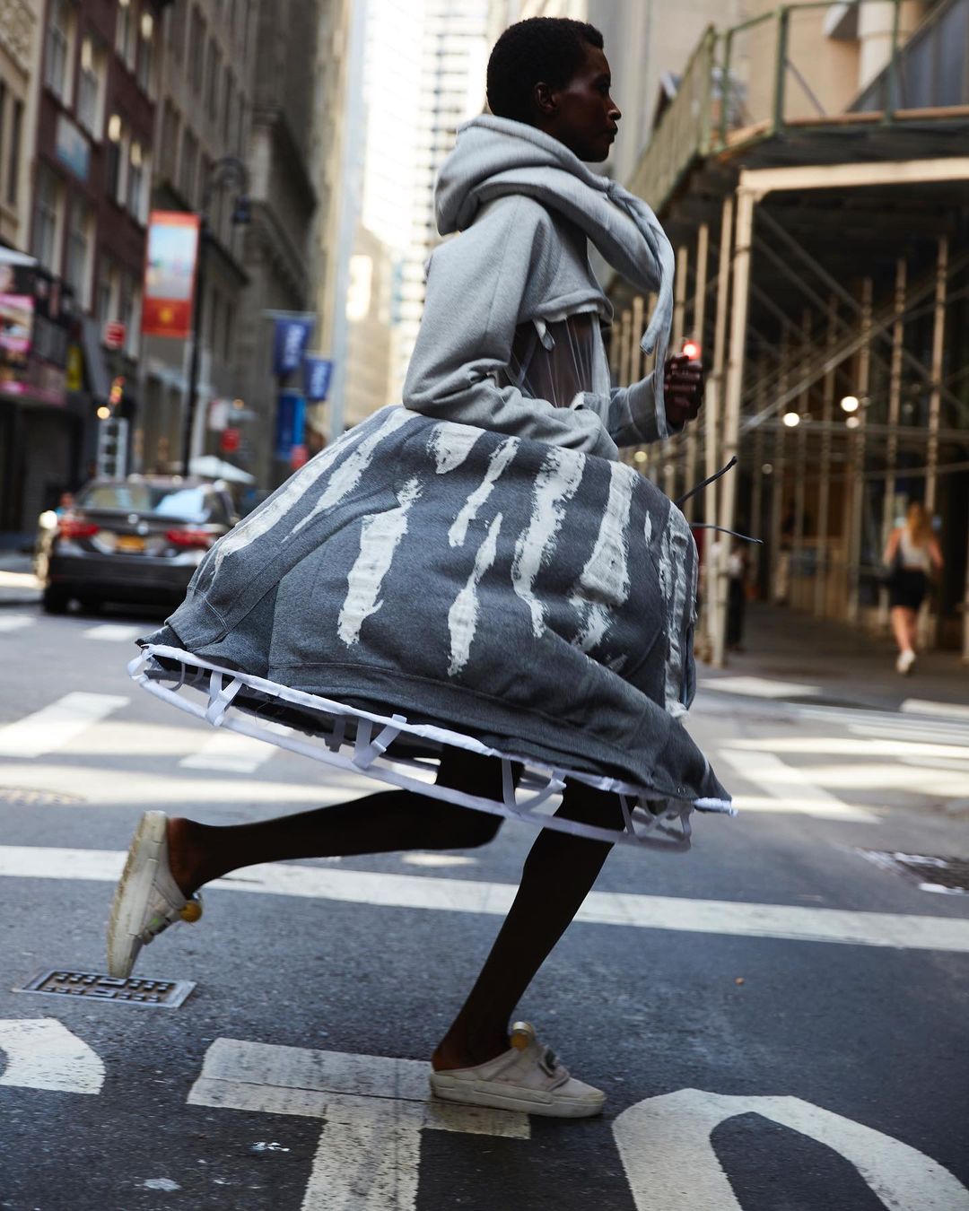 A person in a grey sculptural dress crosses a city street, captured in motion with buildings and a car in the background.