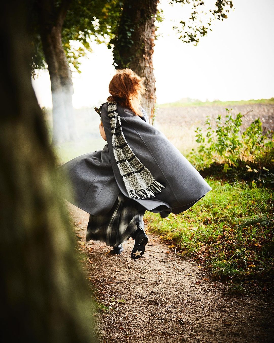 A woman in a grey cape and plaid skirt walks on a dirt path lined with trees in a blurred outdoor setting, viewed from behind.