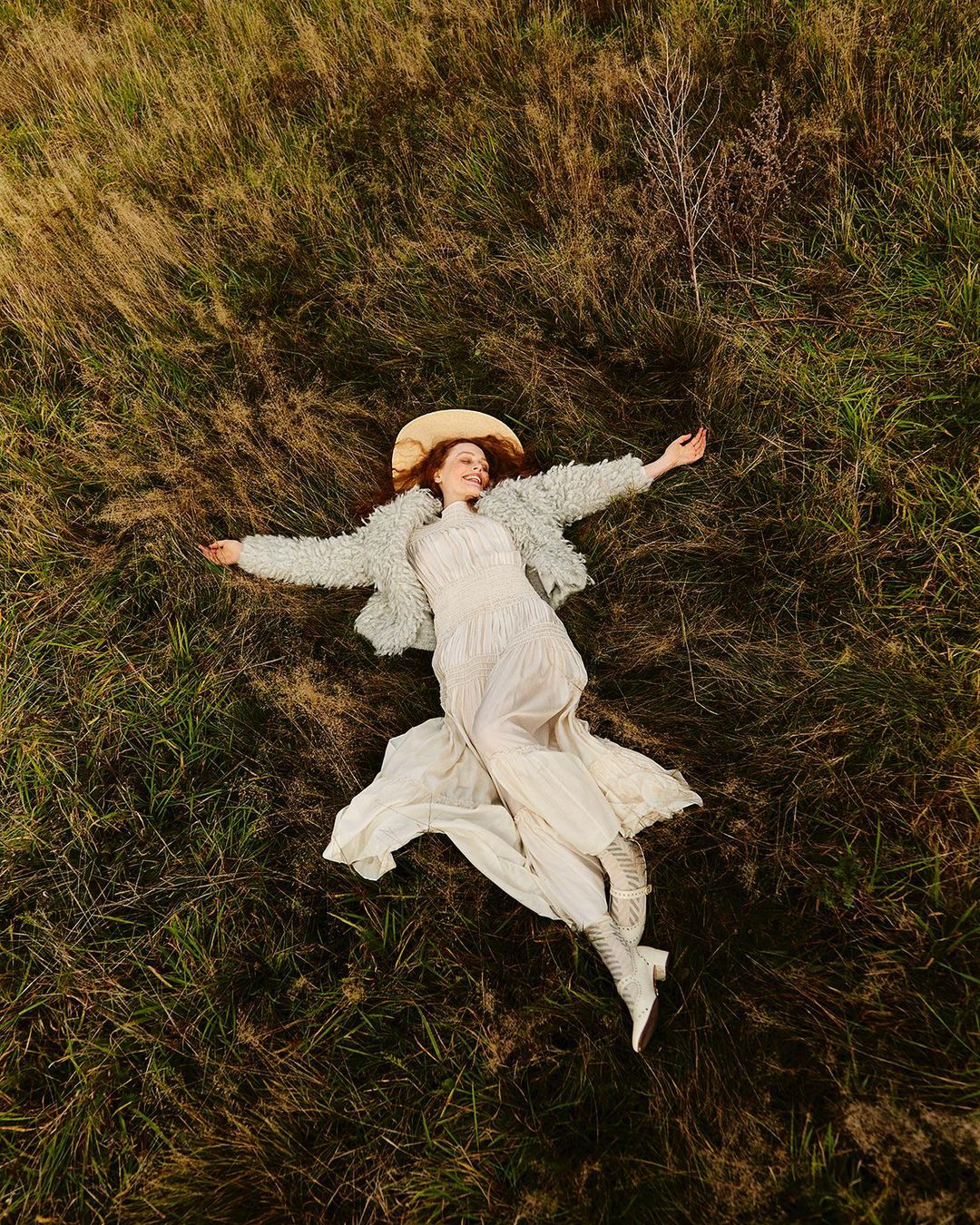 Overhead view of a woman lying in tall grass wearing a white dress and textured jacket with a hat