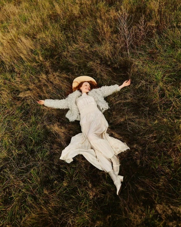 Overhead view of a woman lying in tall grass wearing a white dress and textured jacket with a hat
