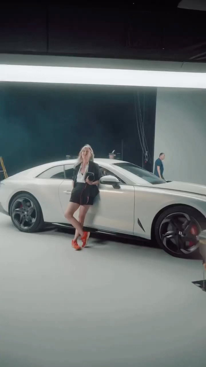 Woman poses with a silver coupe in a photo studio setting; a car photoshoot with artificial lighting and light gray backdrop.