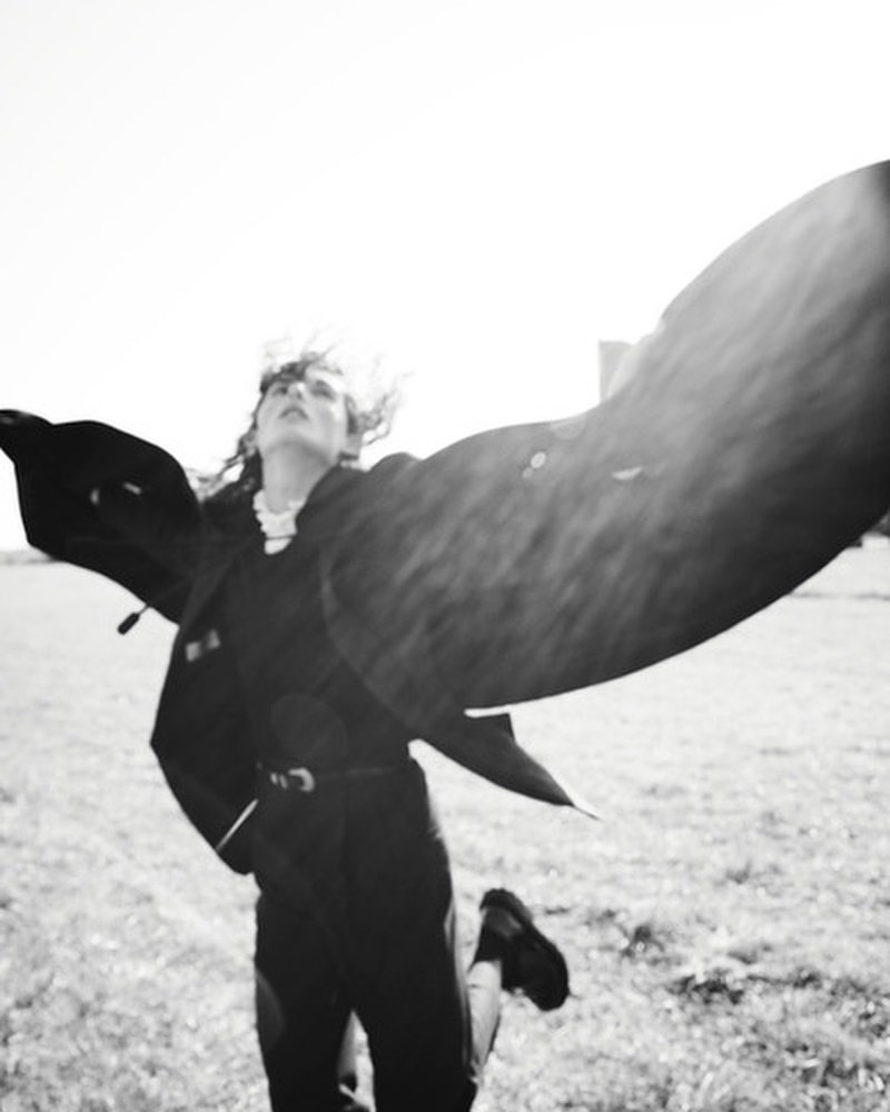 Black-and-white outdoor shot of a woman with tousled hair and a jacket with open arms, moving in a grassy field under bright sunlight.