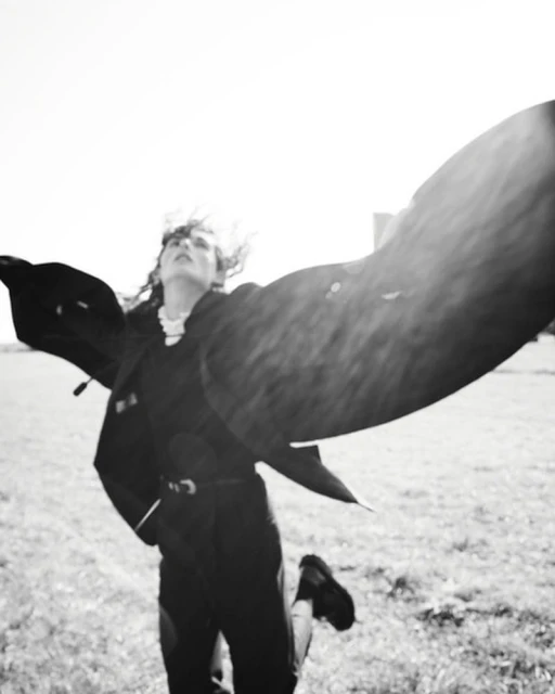 Black-and-white outdoor shot of a woman with tousled hair and a jacket with open arms, moving in a grassy field under bright sunlight.