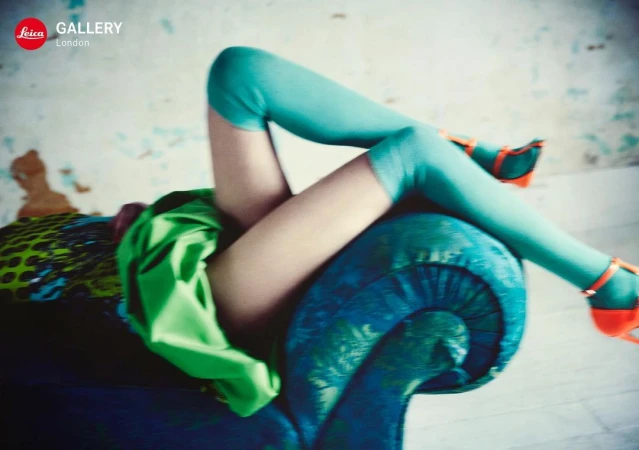 Woman lounges on a blue chaise, showcasing turquoise hosiery and orange heels against a textured wall, with a bold green garment draped nearby.