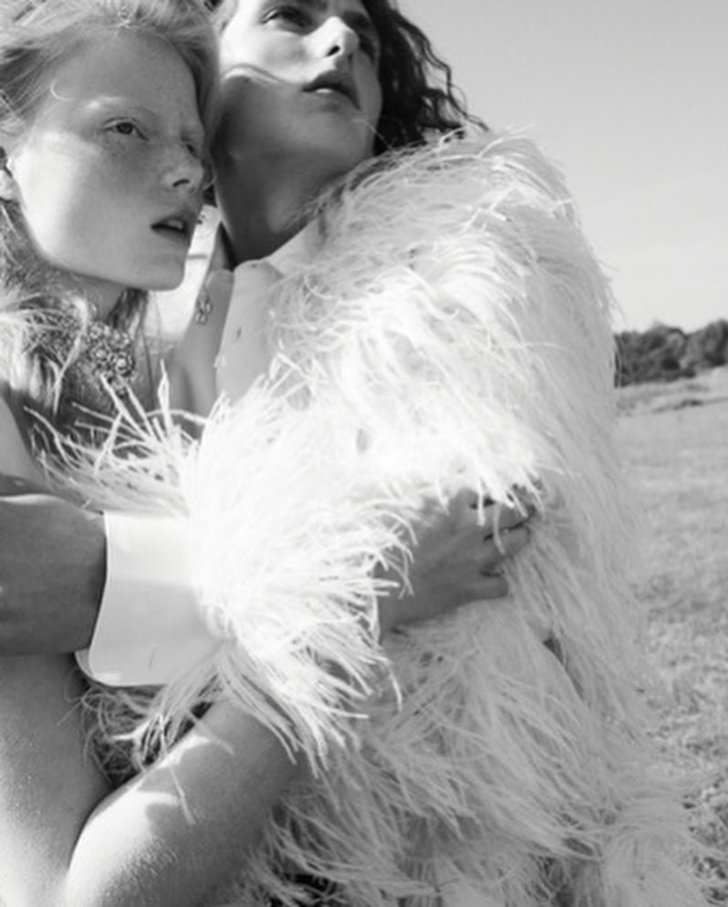 Two women in a black and white fashion shot. One woman is wrapped in a white, feathered jacket in an outdoor field setting.