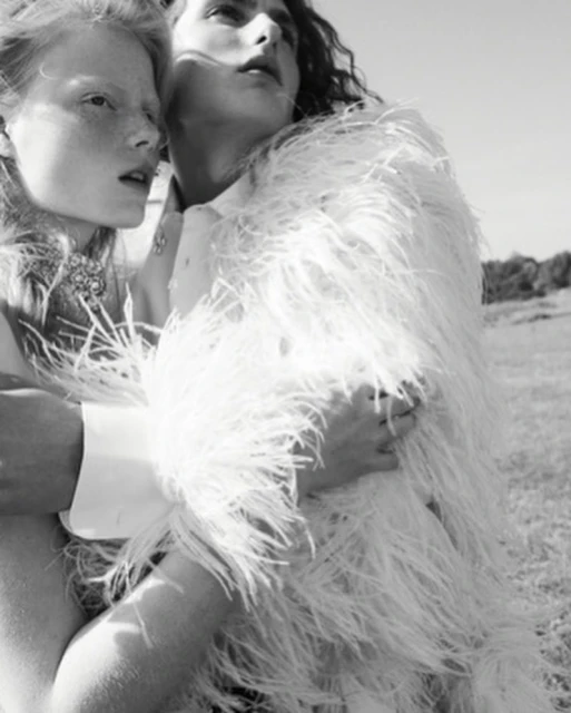 Two women in a black and white fashion shot. One woman is wrapped in a white, feathered jacket in an outdoor field setting.