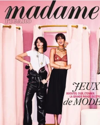 Two female models posing in fitting rooms with pink curtains, one wearing black leather pants and a white t-shirt, the other in a red lace bralette.