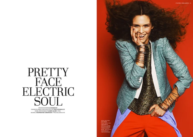 Smiling model with voluminous dark curly hair wearing a blue tweed jacket and stacked silver bangles against a red background