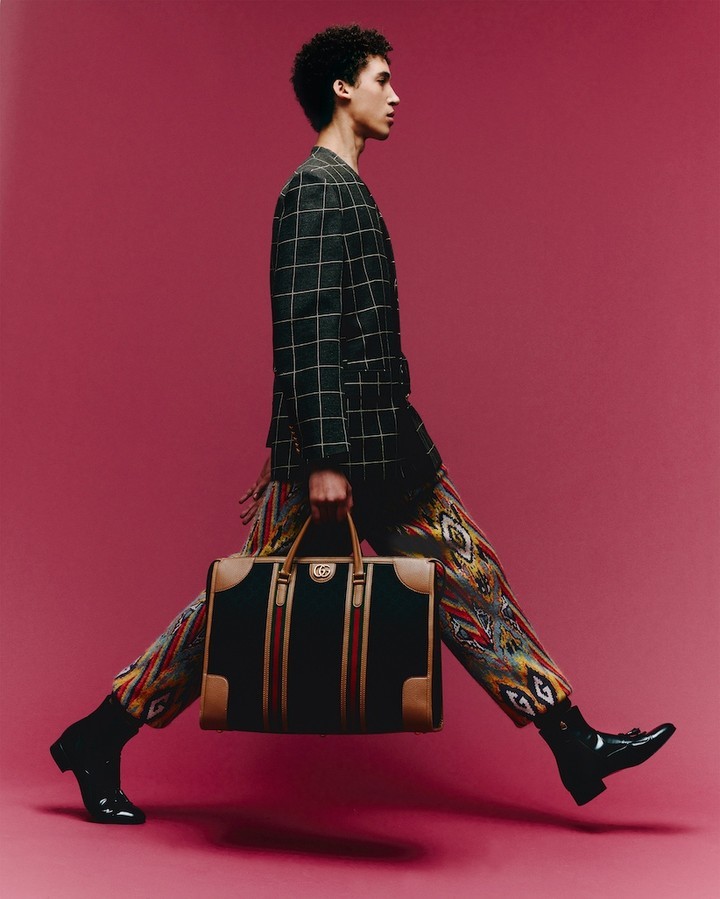 Fashion shot of a young adult male walking and carrying a Gucci travel bag in a studio with a pink background and wearing a suit.