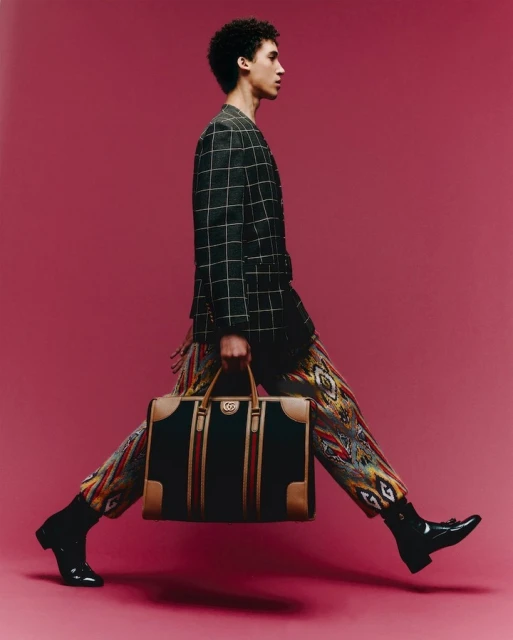Fashion shot of a young adult male walking and carrying a Gucci travel bag in a studio with a pink background and wearing a suit.
