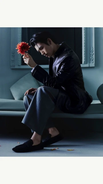 A man in a blazer and trousers sits on a couch holding a red flower, showcasing fashion and style in an indoor setting.
