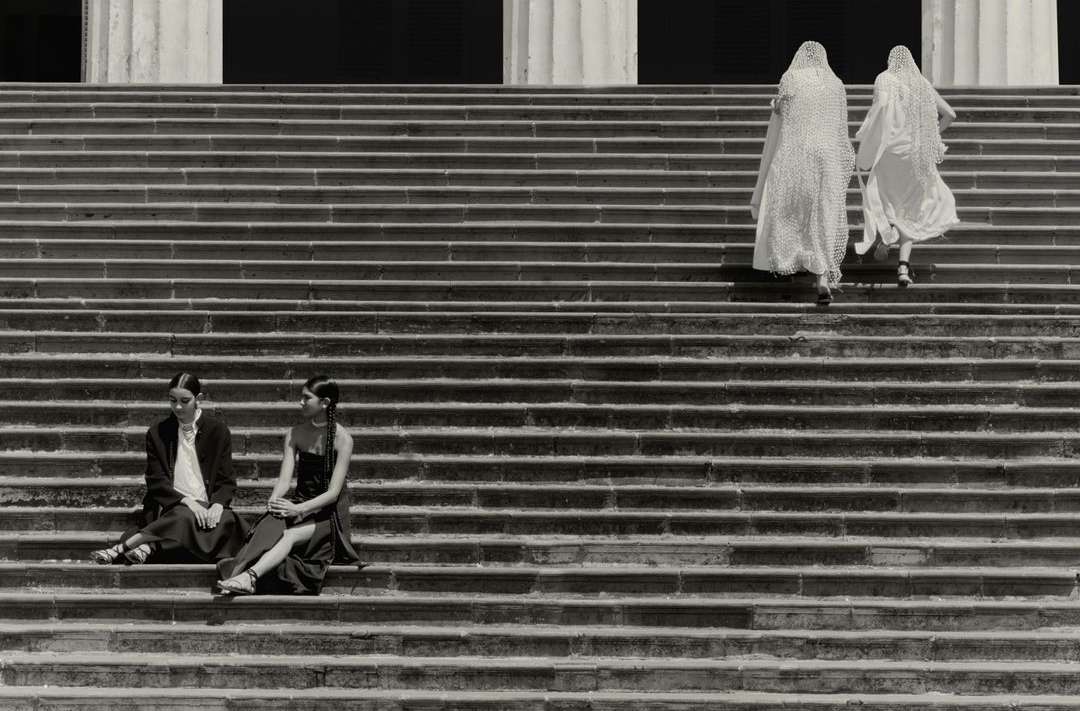 Two women in dark dresses sit on stone steps while two women in white climb steps in a black and white architectural outdoor scene.