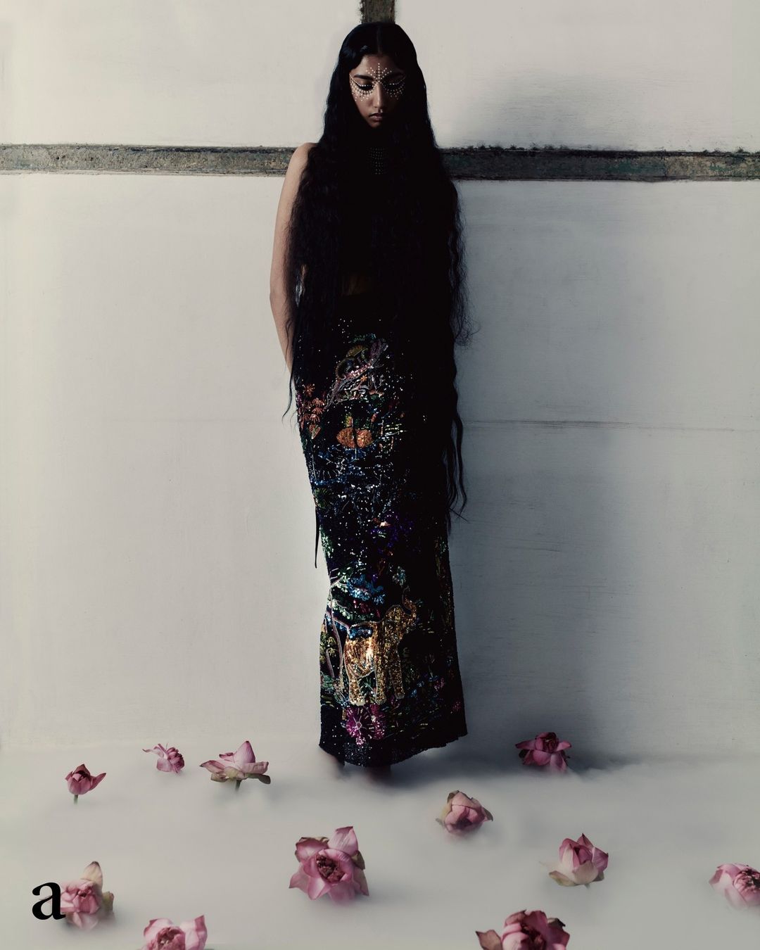 Woman with long dark hair in a patterned dress standing indoors among scattered pink roses in a fine art portrait.