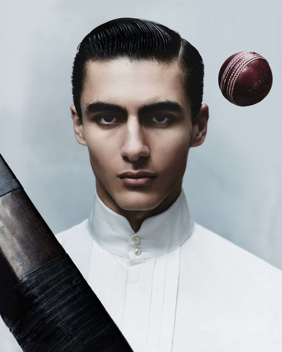 Portrait of a handsome adult male in formal white shirt holding cricket bat with cricket ball in background, posing in a studio.