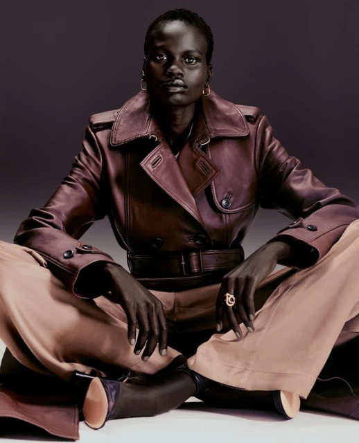 Fashion portrait of a Black woman in a leather trench coat, seated on the floor with tan pants and dark boots in a grey studio.
