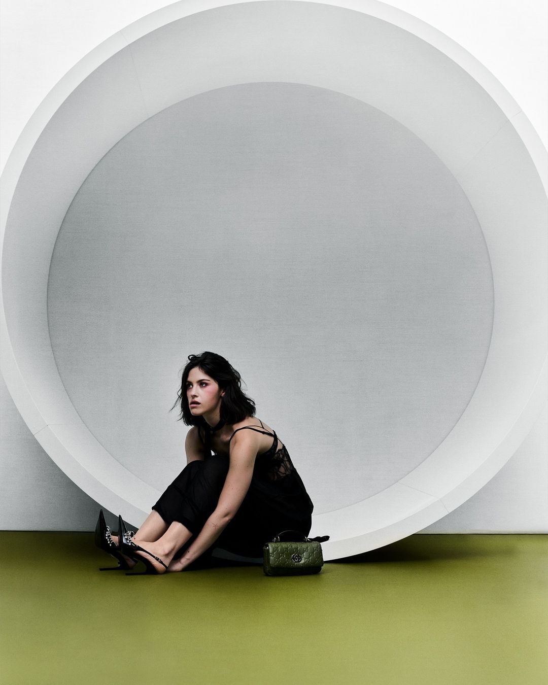 Woman in black outfit sits in front of white circular structure in a studio setting with minimalist style and a small green purse.