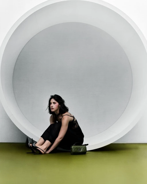 Woman in black outfit sits in front of white circular structure in a studio setting with minimalist style and a small green purse.