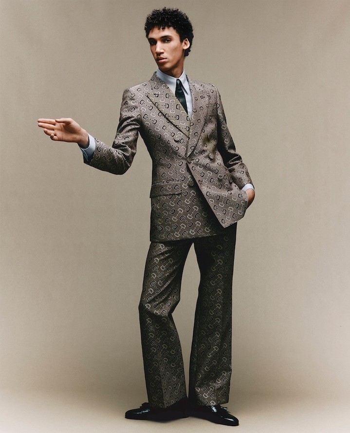 Man wearing a patterned brown suit and tie gesturing forward against a seamless neutral grey studio background