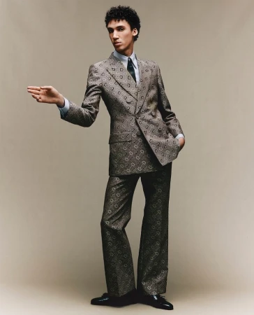 Man wearing a patterned brown suit and tie gesturing forward against a seamless neutral grey studio background