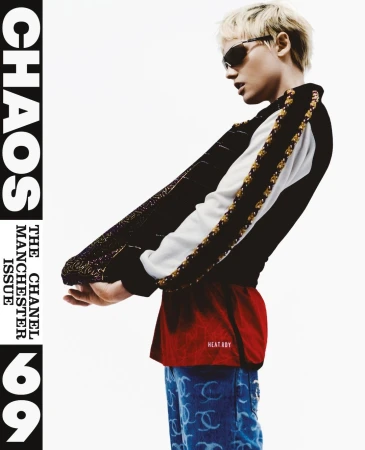Model with blonde hair and sunglasses wearing a black jacket, red shirt, and patterned jeans against a plain background