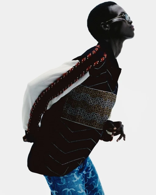 Fashion shot of a dark-skinned adult model wearing sunglasses, blue logo-patterned jeans, and a patterned jacket on a bright backdrop.