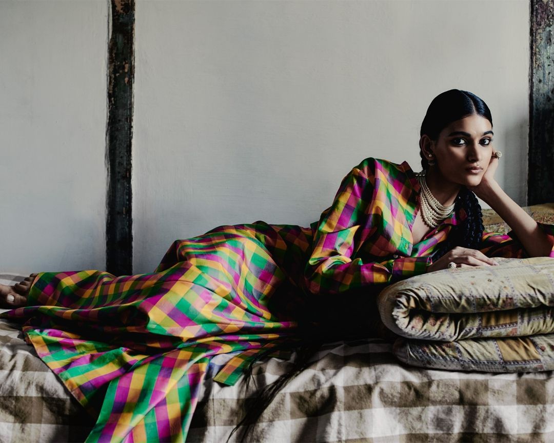 Adult female model poses on a bed in an indoor setting, wearing a colorful plaid dress, layered pearl necklace, and earrings.