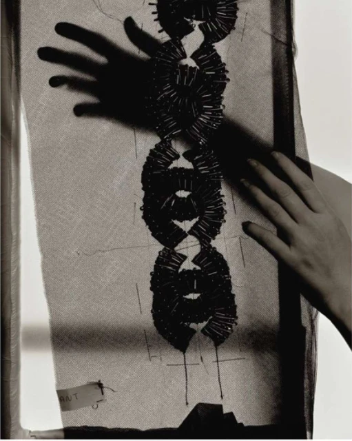 Close-up of a sheer fabric with circular black beaded embellishments, hand visible, creating shadows. Monochromatic color palette.