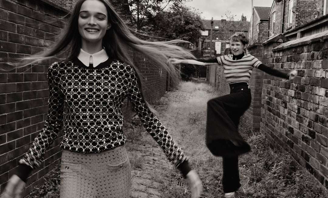 Two happy women in black-and-white fashion photograph. One long-haired woman smiles, while the other leaps by a brick wall on a city street.