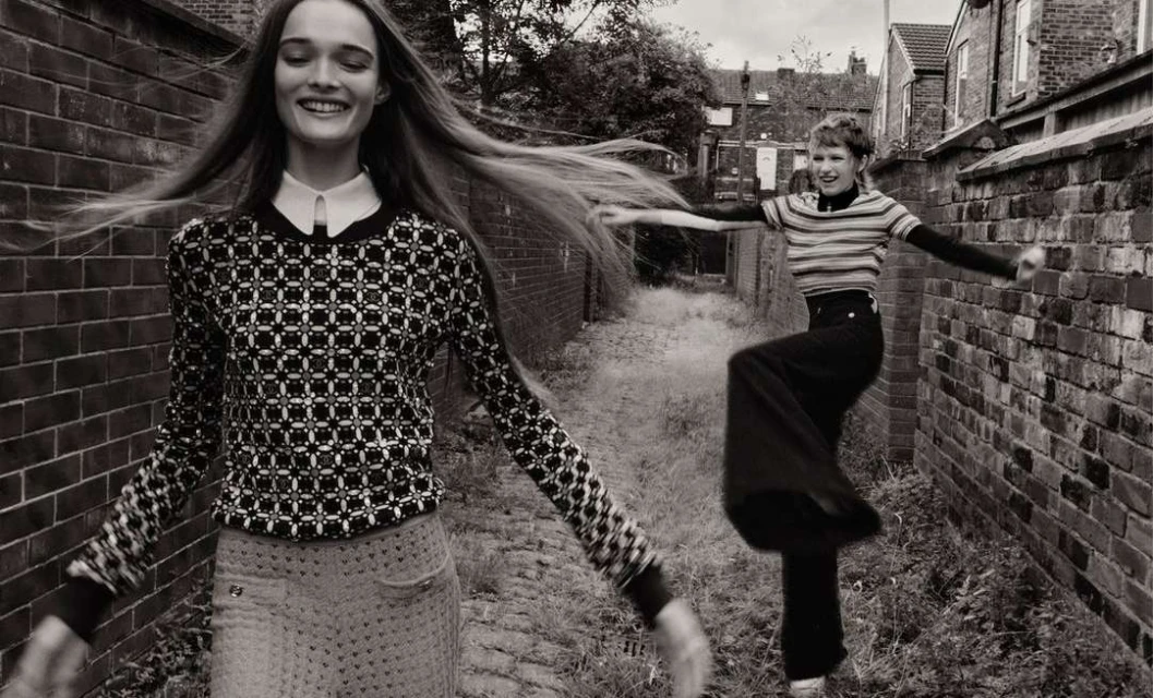 Two happy women in black-and-white fashion photograph. One long-haired woman smiles, while the other leaps by a brick wall on a city street.
