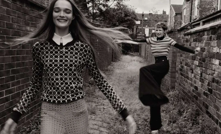 Two happy women in black-and-white fashion photograph. One long-haired woman smiles, while the other leaps by a brick wall on a city street.