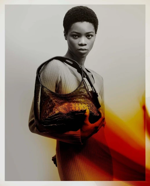 Woman presents an alligator handbag in a fashion editorial style, wearing neutral clothing with dramatic lighting against a simple backdrop.