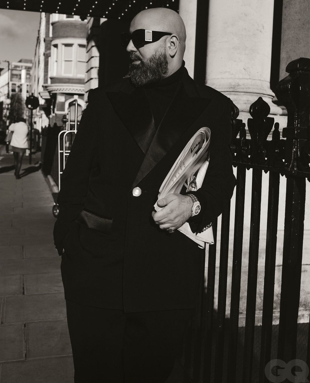 Monochrome portrait of a bald adult male in sunglasses and a dark jacket, holding magazines against an urban backdrop.