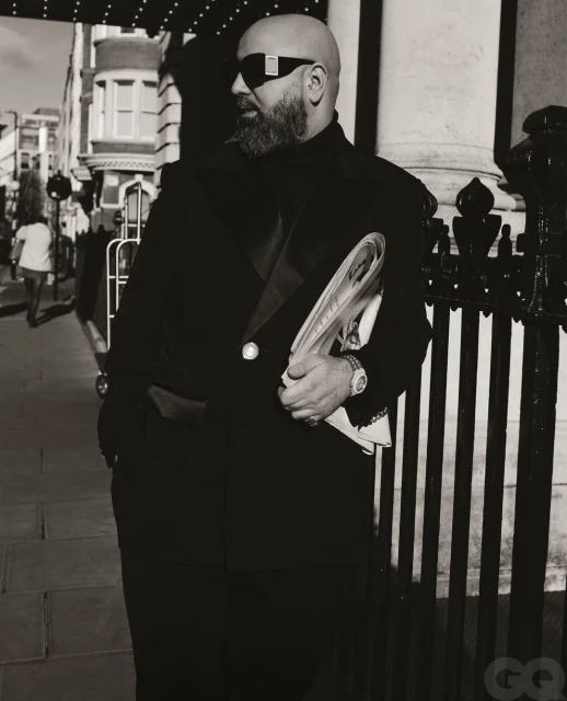 Monochrome portrait of a bald adult male in sunglasses and a dark jacket, holding magazines against an urban backdrop.