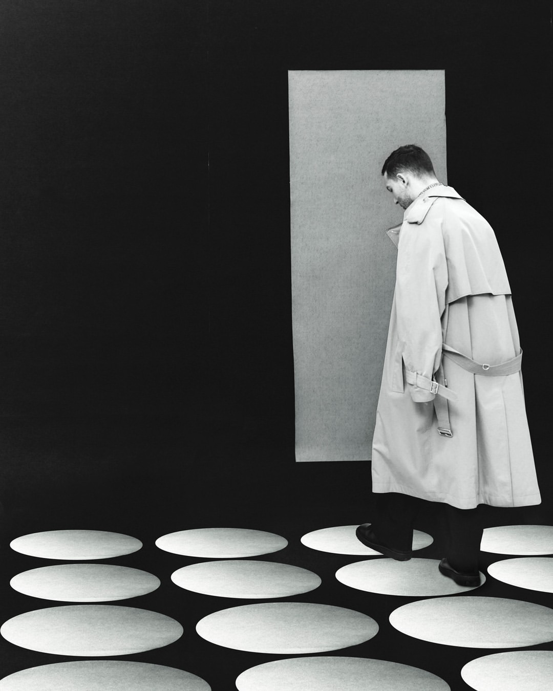 A man in a light beige trench coat walks on white oval shapes over a black surface, in front of a light gray rectangular panel.