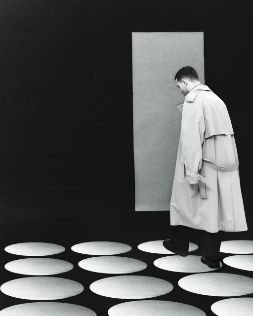 A man in a light beige trench coat walks on white oval shapes over a black surface, in front of a light gray rectangular panel.