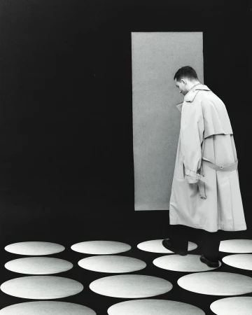 A man in a light beige trench coat walks on white oval shapes over a black surface, in front of a light gray rectangular panel.