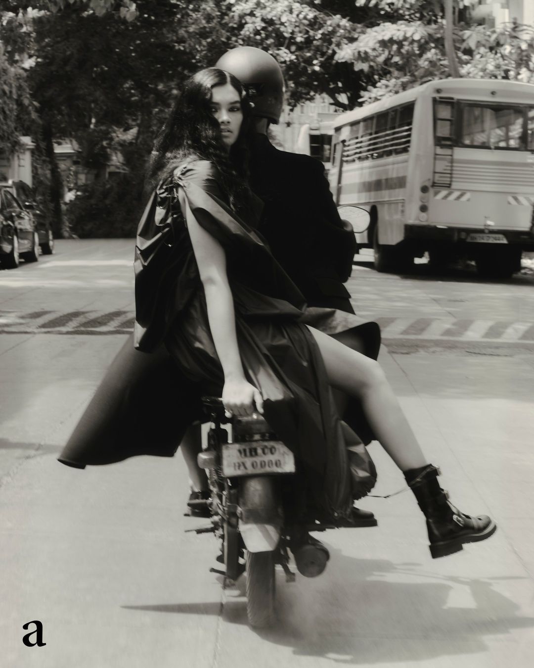 A woman in a dress sits on a vintage motorcycle with a helmeted driver in a black and white street scene. The motorcycle is on a city street.