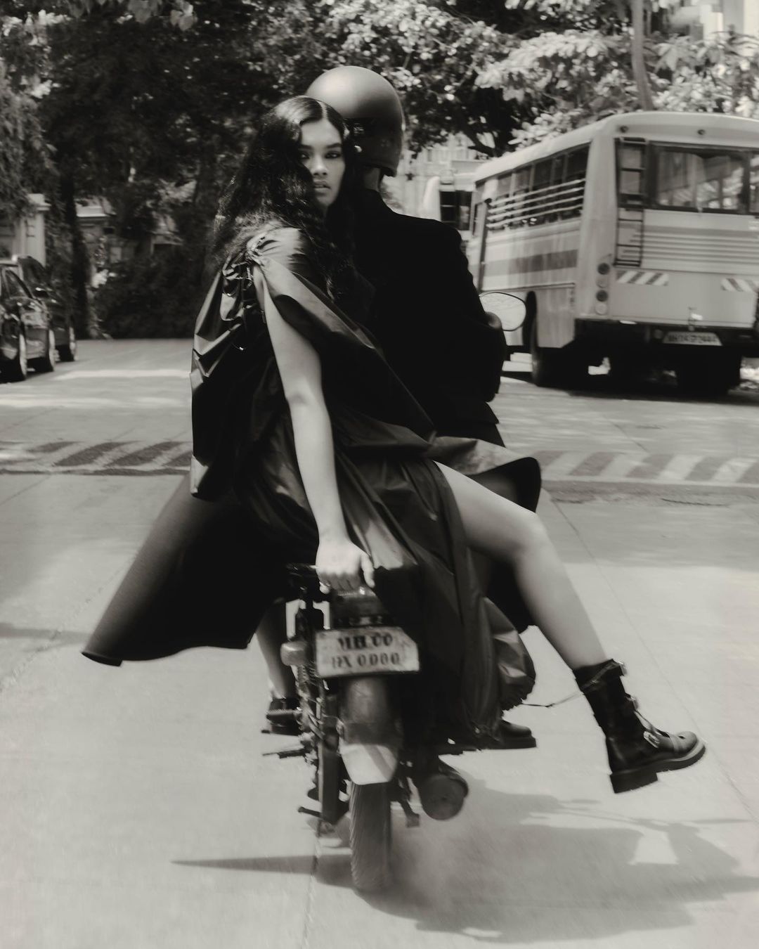 Monochrome editorial photo of a woman in a dark coat riding on the back of a motorcycle on a city street