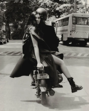 Monochrome editorial photo of a woman in a dark coat riding on the back of a motorcycle on a city street