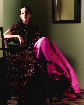 Woman in sequined burgundy dress and bright pink skirt seated in a dark chair against a green wall