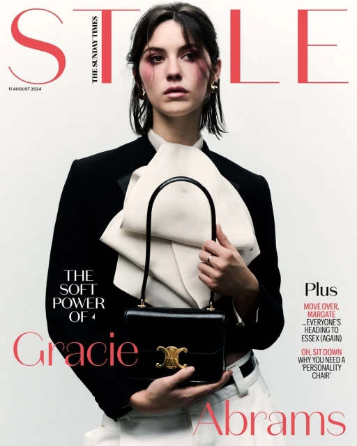Gracie Abrams on magazine cover in blazer holding black purse, photographed indoors with bright, neutral studio lighting and soft focus.