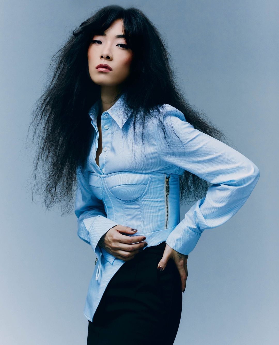 A stylish Asian woman with dark hair poses in a studio wearing a light blue corset top and black skirt against a light grey background.