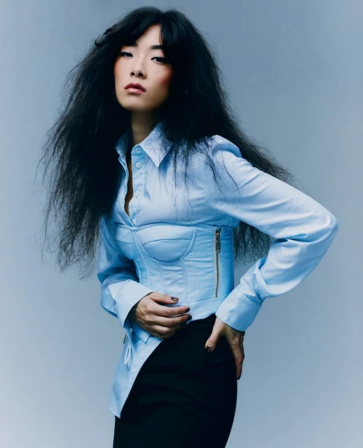 A stylish Asian woman with dark hair poses in a studio wearing a light blue corset top and black skirt against a light grey background.