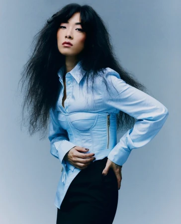 A stylish Asian woman with dark hair poses in a studio wearing a light blue corset top and black skirt against a light grey background.