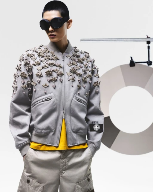 Asian model in a studio wearing a gray bomber jacket with floral appliques, yellow shirt, khaki shorts, and black sunglasses with the Dior logo.