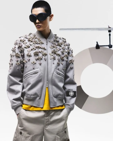 Asian model in a studio wearing a gray bomber jacket with floral appliques, yellow shirt, khaki shorts, and black sunglasses with the Dior logo.